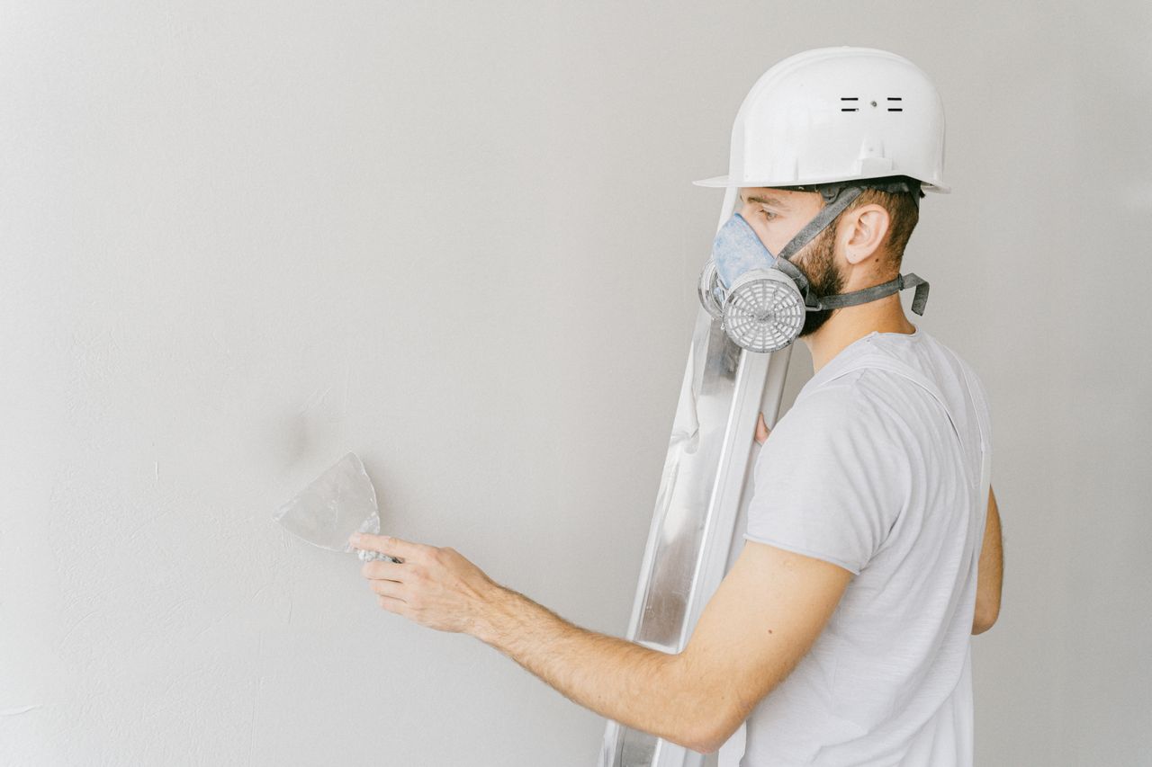 A construction worker wearing a gas mask and hard hat applies plaster to a wall indoors.